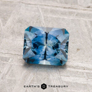 A 1.27-carat radiant-cut blue Montana sapphire (heated) with geometric facets is displayed on a textured gray surface, with the "EARTH'S TREASURY" text and logo below.
