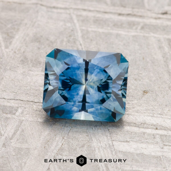 1.27-Carat Montana Sapphire (Heated) A 1.27-carat radiant-cut blue Montana sapphire (heated) with geometric facets is displayed on a textured gray surface, with the "EARTH'S TREASURY" text and logo below.