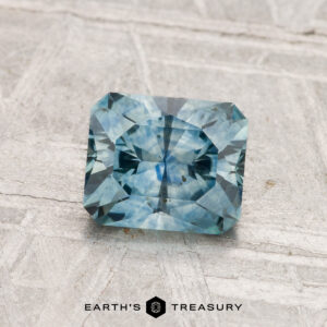 A 1.26-carat heated Montana sapphire with a rectangular blue-green facet is displayed on a light textured surface, with "EARTH'S TREASURY" written below.