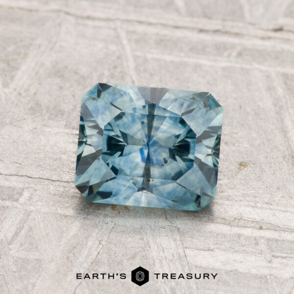 1.26-Carat Montana Sapphire (Heated) A 1.26-carat heated Montana sapphire with a rectangular blue-green facet is displayed on a light textured surface, with "EARTH'S TREASURY" written below.