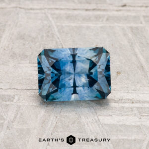 A 1.11-carat rectangular blue Montana sapphire (heated) with angular facets rests on a light textured surface. "EARTH'S TREASURY" appears at the bottom.