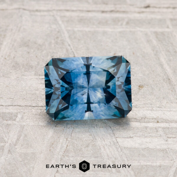 1.11-Carat Montana Sapphire (Heated) A 1.11-carat rectangular blue Montana sapphire (heated) with angular facets rests on a light textured surface. "EARTH'S TREASURY" appears at the bottom.