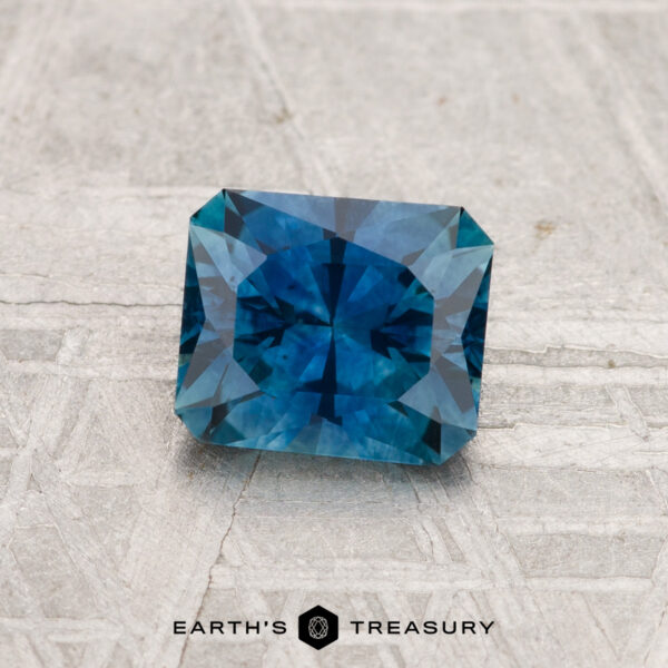 1.10-Carat Montana Sapphire (Heated) A 1.10-carat heated Montana sapphire with a blue, rectangular step-cut is displayed on a light textured surface. The text “EARTH'S TREASURY” appears at the bottom.