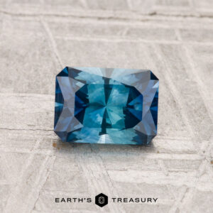 A 1.08-carat heated Montana sapphire with faceted edges is displayed on a textured gray surface, with "EARTH'S TREASURY" below.