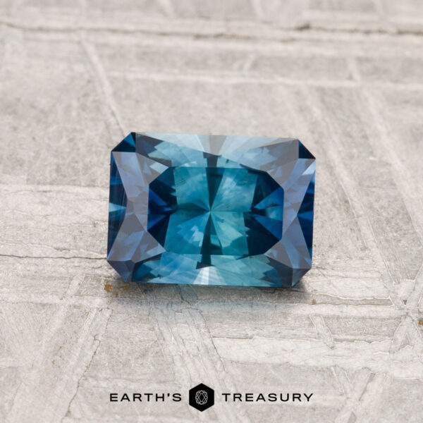 1.08-Carat Montana Sapphire (Heated) A 1.08-carat heated Montana sapphire with faceted edges is displayed on a textured gray surface, with "EARTH'S TREASURY" below.