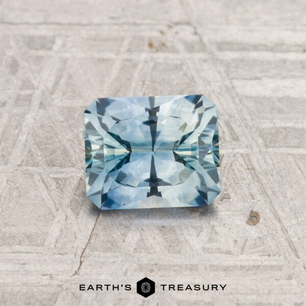 0.83-Carat Montana Sapphire (Heated) A rectangular, faceted 0.83-carat heated Montana sapphire with a blue-green hue is displayed on a textured light gray surface above the text "EARTH'S TREASURY.