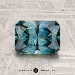 A rectangular step-cut 2.70-carat heated Montana sapphire in blue-green is displayed on a light textured surface, with "EARTH'S TREASURY" at the bottom.