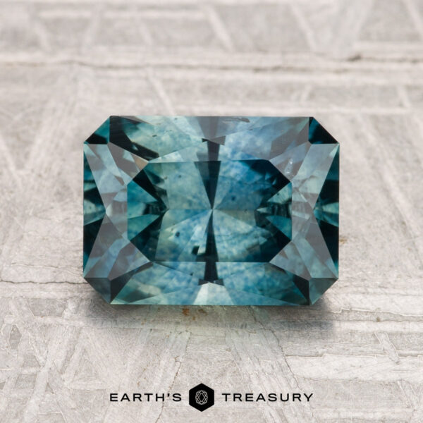 2.70-Carat Montana Sapphire (Heated) A rectangular step-cut 2.70-carat heated Montana sapphire in blue-green is displayed on a light textured surface, with "EARTH'S TREASURY" at the bottom.
