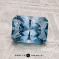 A 2.54-carat heated Montana sapphire with a radiant cut is displayed on a textured light gray surface. The "EARTH'S TREASURY" logo appears at the bottom of the image.