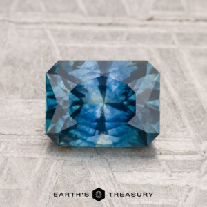 A 1.82-carat heated Montana sapphire, cut in a blue emerald shape with geometric facets, rests on a textured, light surface. "EARTH'S TREASURY" appears at the bottom.