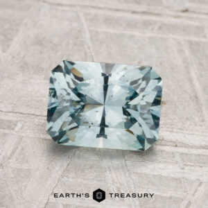 A 1.73-carat rectangular, faceted Montana Sapphire (heated) with a light blue hue is shown on a textured pale surface, with "Earth's Treasury" displayed below.