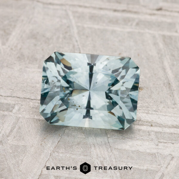 1.73-Carat Montana Sapphire (Heated) A 1.73-carat rectangular, faceted Montana Sapphire (heated) with a light blue hue is shown on a textured pale surface, with "Earth's Treasury" displayed below.