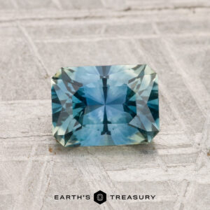 A 1.29-carat heated Montana sapphire, rectangular and faceted in blue-green hues, showcased on a light textured surface with "EARTH'S TREASURY" beneath.