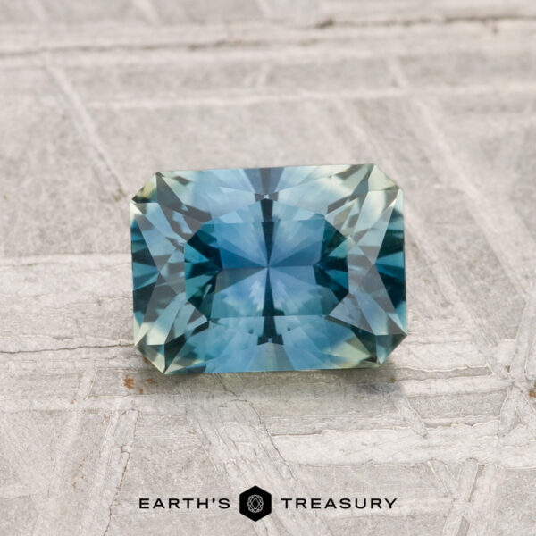 1.29-Carat Montana Sapphire (Heated) A 1.29-carat heated Montana sapphire, rectangular and faceted in blue-green hues, showcased on a light textured surface with "EARTH'S TREASURY" beneath.
