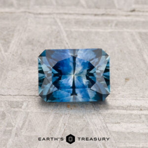 A 1.10-carat heated Montana sapphire with a geometric rectangular cut rests on a textured gray surface. The Earth's Treasury logo is at the bottom of the image.