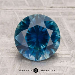 A 3.05-carat heated Montana sapphire with a round brilliant cut is displayed on a textured, light-colored surface with the text "EARTH'S TREASURY" below.