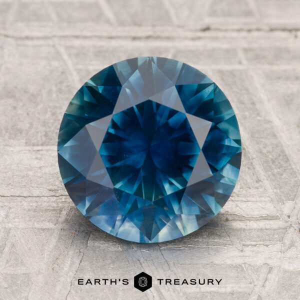 3.05-Carat Montana Sapphire (Heated) A 3.05-carat heated Montana sapphire with a round brilliant cut is displayed on a textured, light-colored surface with the text "EARTH'S TREASURY" below.