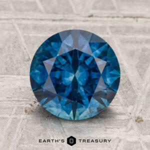 A 2.63-carat heated Montana sapphire, round and faceted in blue, is shown on a textured light surface with the Earth's Treasury logo below.