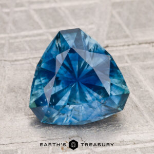 A faceted, triangular 2.86-carat heated Montana sapphire rests on a textured gray surface with the "Earth's Treasury" logo at the bottom.