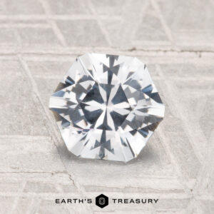 A faceted, octagonal 1.49-carat Montana Sapphire rests on a textured light gray surface with the "Earth's Treasury" logo and text at the bottom.