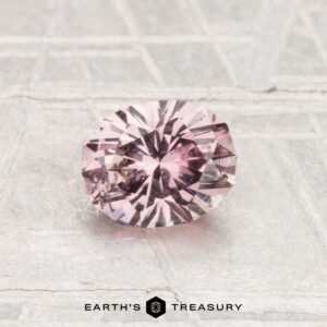 A 0.86-carat Montana sapphire, faceted in an oval shape with a pink hue, is displayed on a textured light-gray surface with "EARTH'S TREASURY" at the bottom.