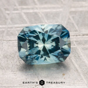 A 2.24-carat heated Montana sapphire with a rectangular faceted cut in blue-green is shown on a textured gray surface, with the "Earth's Treasury" logo below.