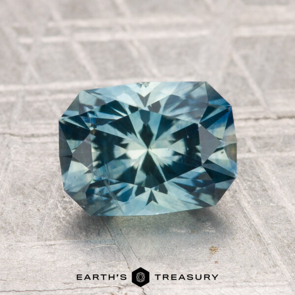 2.24-Carat Montana Sapphire (Heated) A 2.24-carat heated Montana sapphire with a rectangular faceted cut in blue-green is shown on a textured gray surface, with the "Earth's Treasury" logo below.