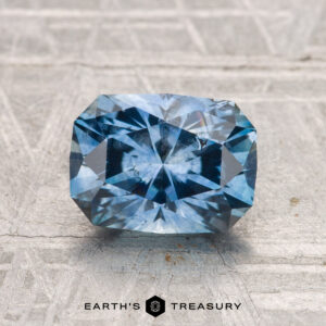 A rectangular, faceted 1.99-carat heated Montana sapphire rests on a gray textured surface with "EARTH'S TREASURY" displayed below.