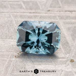 A rectangular, faceted blue 1.73-carat heated Montana sapphire sits on a textured gray surface, with the text "EARTH'S TREASURY" at the bottom.