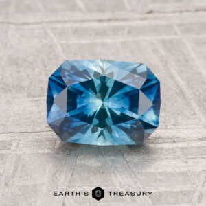 A 1.68-carat heated Montana sapphire with intricate facets lies on a textured gray surface. "EARTH'S TREASURY" is printed at the bottom of the image.