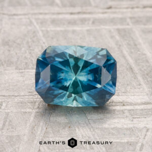 A 1.38-carat heated Montana sapphire, cut in a rectangular blue-green facet, is displayed on a textured light gray surface with "Earth's Treasury" at the bottom.