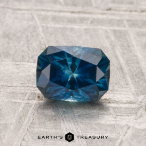 A 1.37-carat heated Montana sapphire with a rectangular, multi-faceted blue-green appearance is displayed on a textured surface, featuring the text "EARTH'S TREASURY" at the bottom.