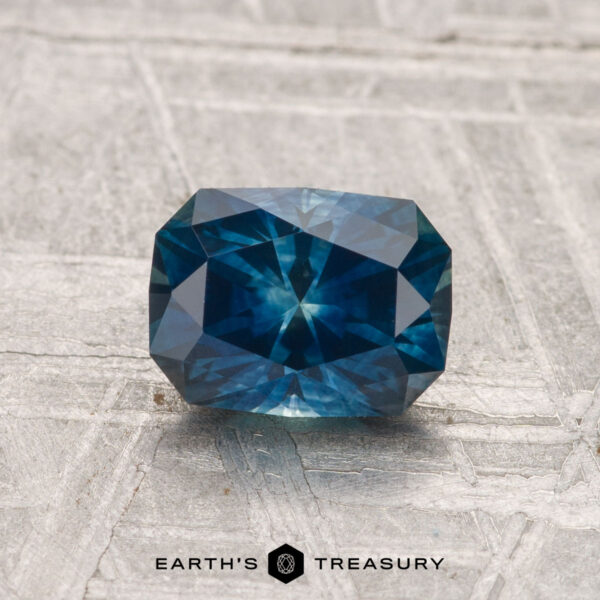 1.37-Carat Montana Sapphire (Heated) A 1.37-carat heated Montana sapphire with a rectangular, multi-faceted blue-green appearance is displayed on a textured surface, featuring the text "EARTH'S TREASURY" at the bottom.