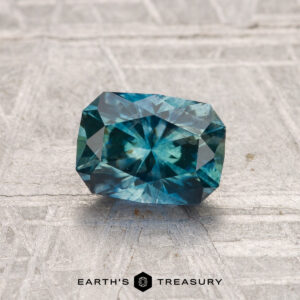 A rectangular, blue-green 1.31-carat heated Montana sapphire rests on a textured surface with "EARTH'S TREASURY" displayed at the bottom.