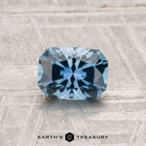 A rectangular, blue 1.02-carat heated Montana sapphire rests on a textured gray surface with "Earth's Treasury" branding at the bottom.