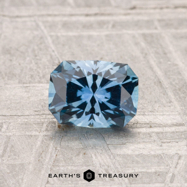 1.02-Carat Montana Sapphire (Heated) A rectangular, blue 1.02-carat heated Montana sapphire rests on a textured gray surface with "Earth's Treasury" branding at the bottom.