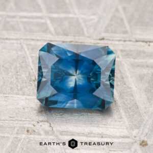 A rectangular, faceted 1.60-carat heated Montana sapphire with a blue hue is shown on a textured grey surface, the "Earth's Treasury" logo displayed below.