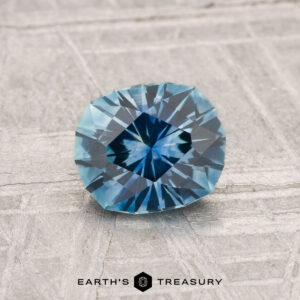 A 1.19-carat heated Montana sapphire, faceted oval and blue, rests on a textured gray surface with "EARTH'S TREASURY" at the bottom.