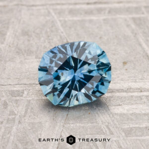 A 1.16-carat heated Montana sapphire with oval radiant cuts is displayed on a textured light gray surface. The Earth's Treasury logo appears at the bottom of the image.
