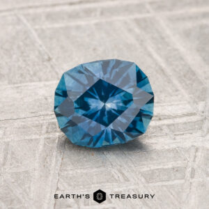 A 1.13-carat heated Montana sapphire, cushion-cut with geometric facets, is displayed on a textured gray surface with the Earth’s Treasury logo at the bottom.