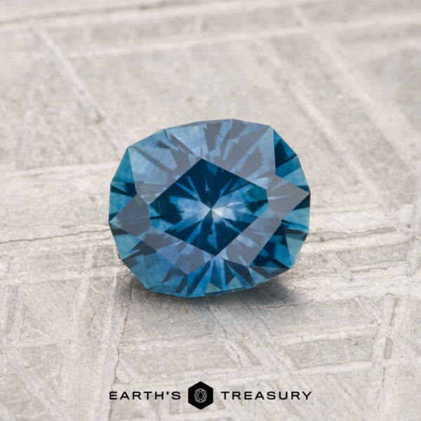 1.13-Carat Montana Sapphire (Heated) A 1.13-carat heated Montana sapphire, cushion-cut with geometric facets, is displayed on a textured gray surface with the Earth’s Treasury logo at the bottom.