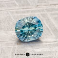 A 1.12-carat heated Montana sapphire with a faceted blue appearance rests on a textured gray surface, with "EARTH'S TREASURY" shown at the bottom of the image.