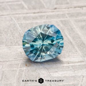 A 1.12-carat heated Montana sapphire with a faceted blue appearance rests on a textured gray surface, with "EARTH'S TREASURY" shown at the bottom of the image.