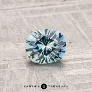 A 0.81-carat heated Montana sapphire, round brilliant-cut in blue-green, is displayed on a textured gray surface with the text "EARTH'S TREASURY" at the bottom.
