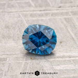 A 0.75-carat heated Montana sapphire, cushion-cut and blue, is displayed on a textured gray surface with "Earth's Treasury" at the bottom.
