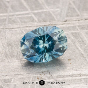 A 1.42-carat heated Montana sapphire is displayed on a textured gray surface with "EARTH'S TREASURY" at the bottom.