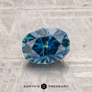 A 1.28-carat heated Montana sapphire with an oval cut is shown on a textured gray surface, with "EARTH'S TREASURY" displayed below.