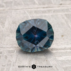 A 1.53-carat heated Montana sapphire with a deep blue hue is shown on a textured gray surface, featuring the text "EARTH'S TREASURY" at the bottom.