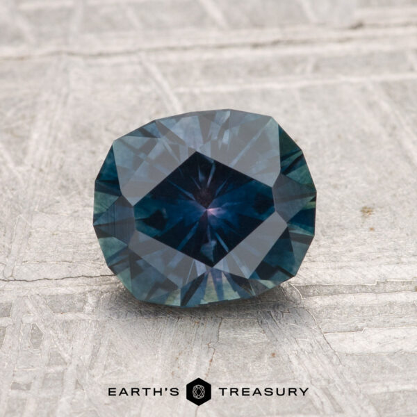 1.53-Carat Montana Sapphire (Heated) A 1.53-carat heated Montana sapphire with a deep blue hue is shown on a textured gray surface, featuring the text "EARTH'S TREASURY" at the bottom.
