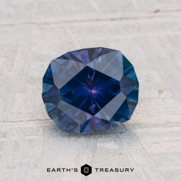 1.53-Carat Montana Sapphire (Heated) A faceted, deep blue 1.53-carat heated Montana Sapphire with an oval cut rests on a light, textured surface. The "EARTH'S TREASURY" logo appears at the bottom of the image.